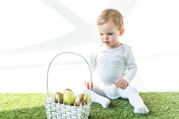 Adorable child sitting on green grass near straw basket with yellow and golden Ester eggs isolated on white — Stock Photo