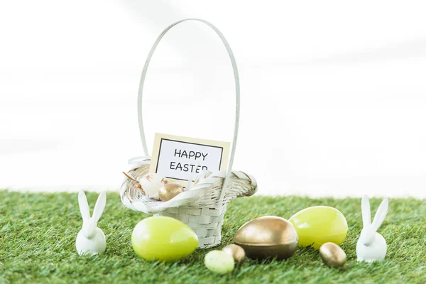 Colorful Easter eggs, decorative rabbits, and straw basket with happy Easter card on green grass isolated on white — Stock Photo