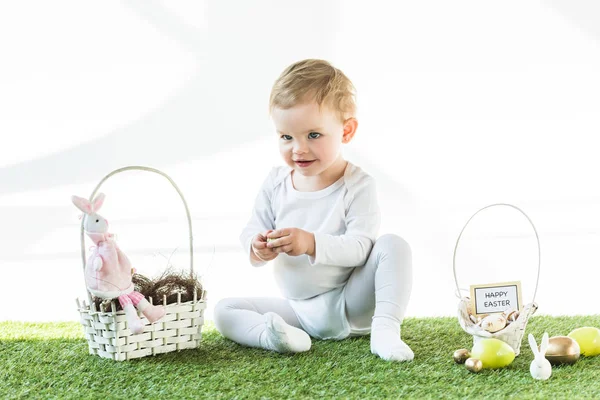 Allegro bambino seduto vicino a cestini di paglia con uova di Pasqua, conigli decorativi e felice carta di Pasqua isolato su bianco — Foto stock