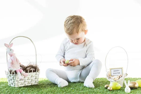Adorable child sitting near straw baskets with Easter eggs, decorative rabbits and happy Easter card isolated on white — Stock Photo