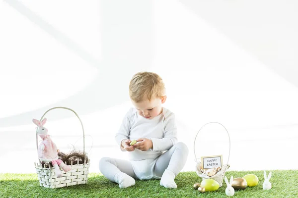 Cute kid sitting near straw baskets with Easter eggs, bunnies and happy Easter card isolated on white — Stock Photo