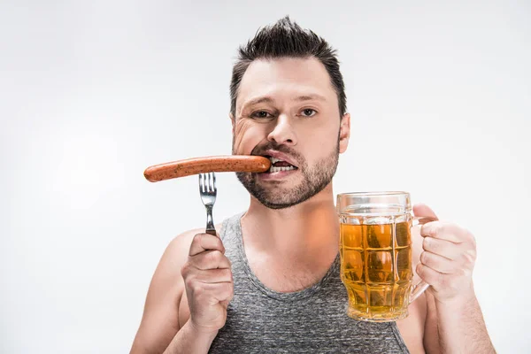 Gordito hombre mordiendo salchicha y sosteniendo vaso de cerveza aislado en blanco - foto de stock
