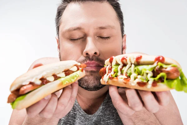 Sobrepeso homem com olhos fechados segurando deliciosos cachorros-quentes isolados em branco — Fotografia de Stock