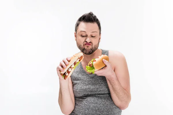 Hombre con sobrepeso sosteniendo perros calientes y haciendo expresión de la cara aislada en blanco - foto de stock