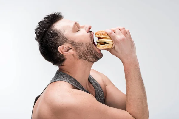 Vista lateral del hombre barbudo gordita comiendo deliciosa hamburguesa aislada en blanco - foto de stock