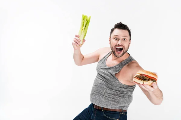 Excited overweight man holding celery and burger isolated on white with copy space — Stock Photo