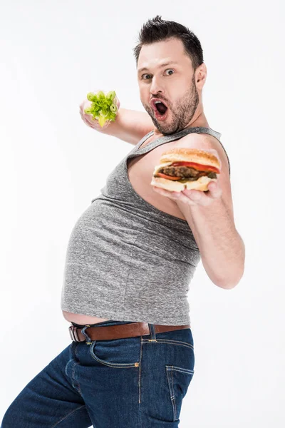 Aufgeregter übergewichtiger Mann blickt in die Kamera, während er Sellerie und Burger isoliert auf Weiß hält — Stockfoto
