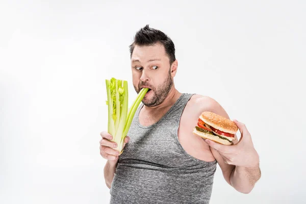 Избыточный вес человек кусает сельдерей, глядя на гамбургер изолирован на белом — стоковое фото