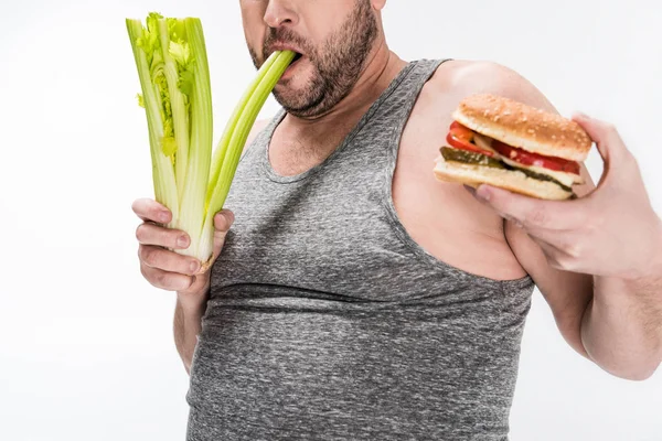 Vista recortada del hombre con sobrepeso mordiendo apio mientras sostiene hamburguesa aislada en blanco - foto de stock