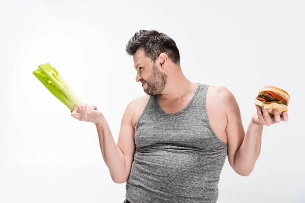Skeptischer übergewichtiger Mann hält Sellerie und Burger isoliert auf weißem Grund — Stockfoto