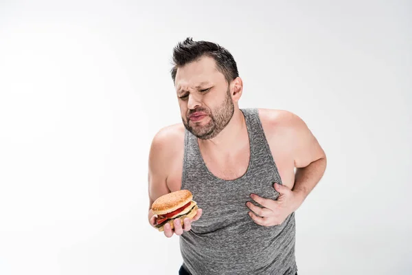 Overweight man holding burger and touching chest on white with copy space — Stock Photo