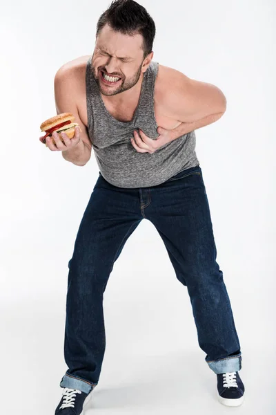 Übergewichtiger Mann im Tanktop hält Burger und berührt Brust auf weißem Grund — Stockfoto