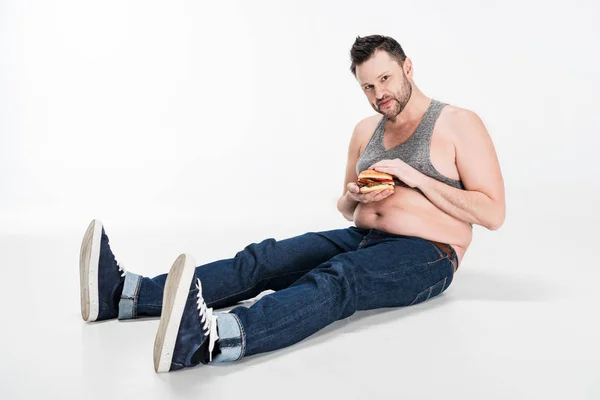 Overweight man sitting with burger and looking at camera on white with copy space — Stock Photo