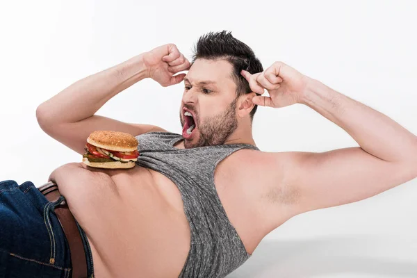 Yelling overweight man with burger on belly lying and doing abs exercise on white — Stock Photo