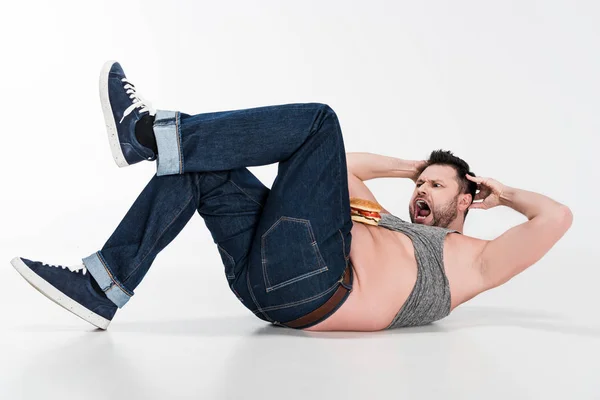 Gritando homem com excesso de peso com hambúrguer na barriga mentindo e fazendo exercícios abdominais em branco — Fotografia de Stock