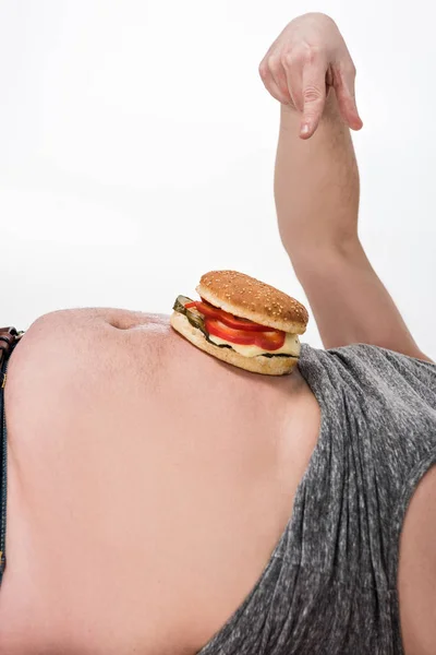 Vue recadrée de l'homme en surpoids pointant avec le doigt au hamburger sur le ventre isolé sur blanc — Photo de stock