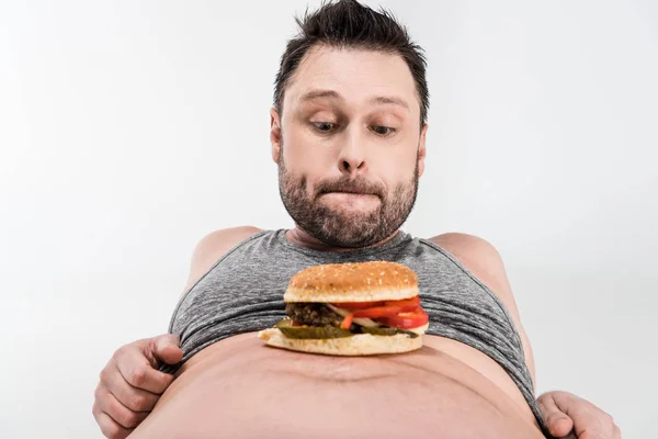 Homme en surpoids regardant hamburger sur le ventre isolé sur blanc — Photo de stock