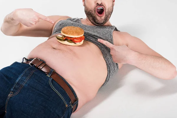 Cropped view of excited overweight man pointing with fingers at burger on belly on white — Stock Photo
