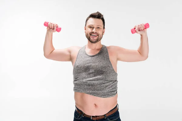 Sonriente hombre con sobrepeso mirando a la cámara mientras hace ejercicio con mancuernas rosadas aisladas en blanco - foto de stock