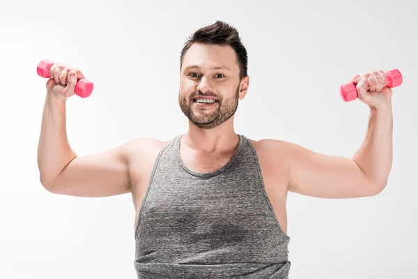 Sonriente hombre con sobrepeso mirando a la cámara mientras hace ejercicio con mancuernas rosadas aisladas en blanco - foto de stock