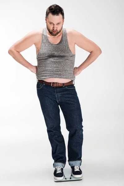 Arrabbiato uomo in sovrappeso con le mani akimbo in piedi su bilance elettroniche di peso e guardando la fotocamera isolata su bianco — Foto stock