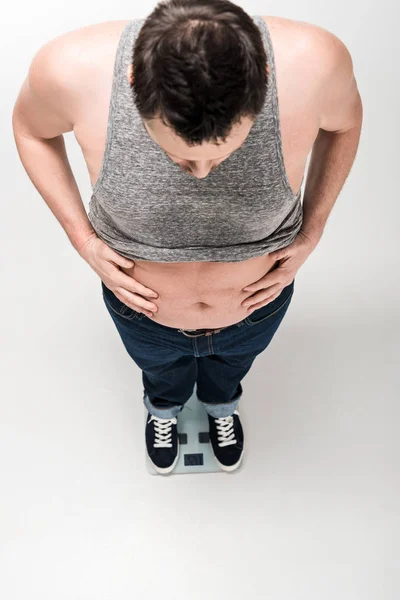 Vista de ángulo alto del hombre con sobrepeso que sostiene el vientre mientras está de pie en balanzas electrónicas aisladas en blanco - foto de stock