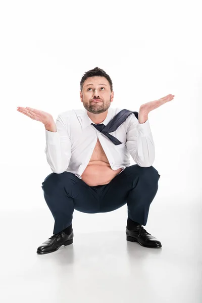 Homem sobrepeso confuso em desgaste formal apertado fazendo gesto de encolher os ombros enquanto sentado no branco — Fotografia de Stock