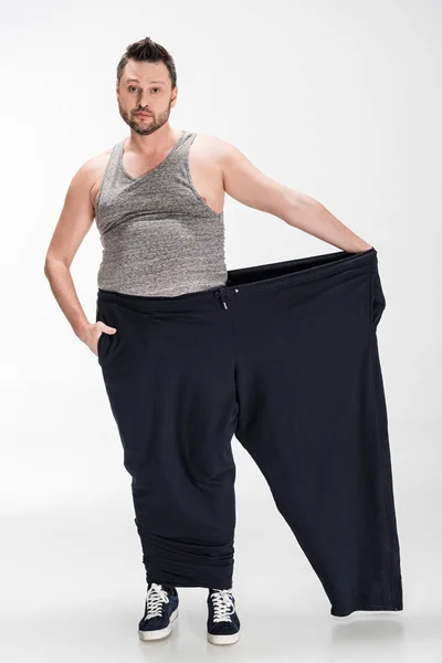 Homme en surpoids tenant pantalon surdimensionné après la perte de poids et en regardant la caméra sur blanc — Photo de stock