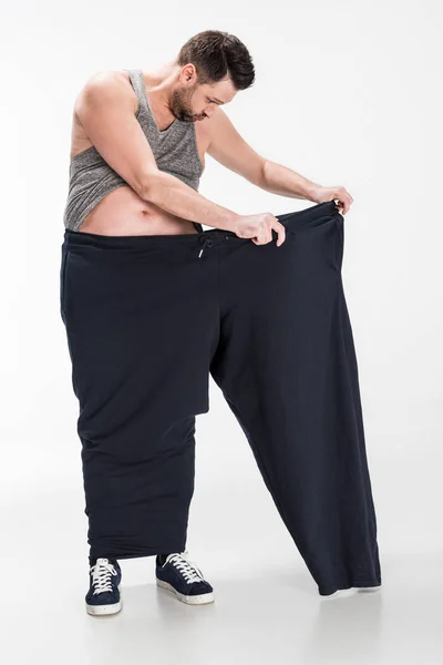 Overweight man in tank top holding oversize pants after weight loss on white — Stock Photo