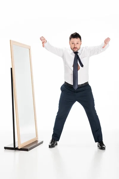 Sovrappeso uomo in abbigliamento formale mostrando pollici verso il basso vicino specchio su bianco — Foto stock