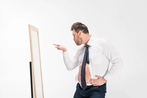 Homem com sobrepeso no desgaste formal apontando com o dedo enquanto olha para o espelho isolado no branco — Fotografia de Stock