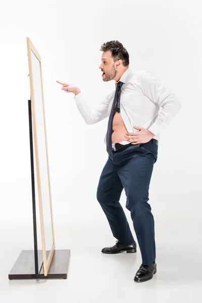 Overweight man in tight formal wear looking at mirror and pointing with finger on white — Stock Photo