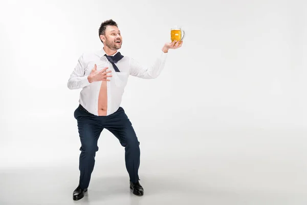 Sonriente hombre con sobrepeso en camisa apretada sosteniendo un vaso de cerveza y haciendo gestos con la mano sobre blanco con espacio para copiar - foto de stock