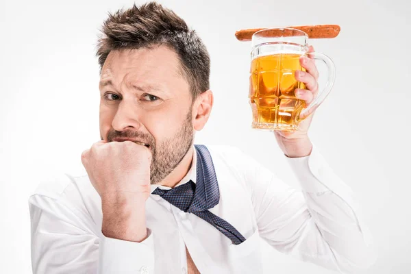 Hombre guapo mordiendo puño y sosteniendo vaso de cerveza con salchicha aislada en blanco - foto de stock