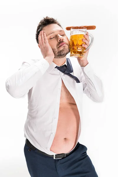 Homem com sobrepeso segurando copo de cerveja com salsicha e posando com os olhos fechados isolado no branco — Fotografia de Stock