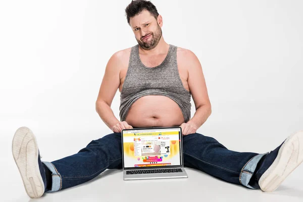 Skeptischer übergewichtiger Mann macht Mimik und sitzt mit Laptop mit aliexpress-Website auf dem Bildschirm isoliert auf weiß — Stockfoto