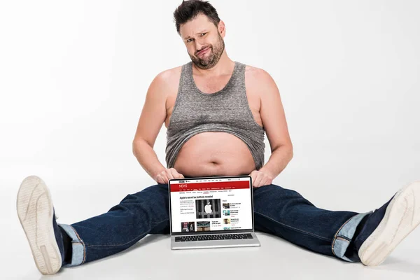 Skeptical overweight man making facial expression and sitting with laptop with bbc news website on screen isolated on white — Stock Photo