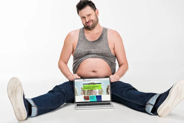 Skeptical overweight man making facial expression and sitting with laptop with bbc website on screen isolated on white — Stock Photo