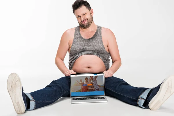 Escéptico hombre con sobrepeso haciendo expresión facial y sentado con el ordenador portátil con couchsurfing sitio web en la pantalla aislado en blanco - foto de stock