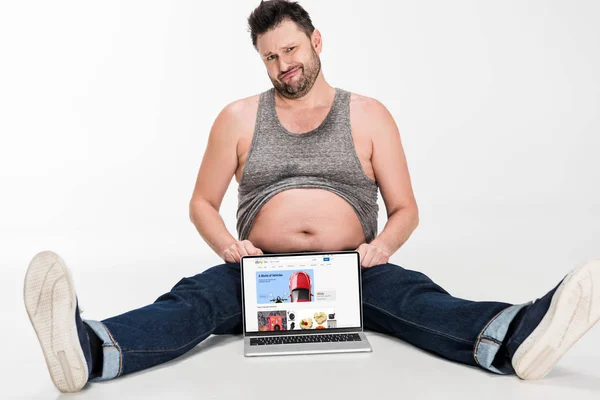 Skeptical overweight man making facial expression and sitting with laptop with ebay website on screen isolated on white — Stock Photo