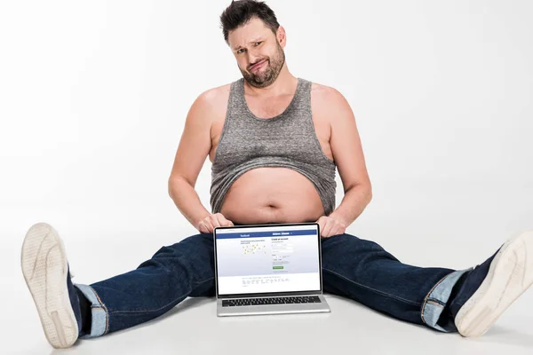 Skeptical overweight man making facial expression and sitting with laptop with facebook website on screen isolated on white — Stock Photo