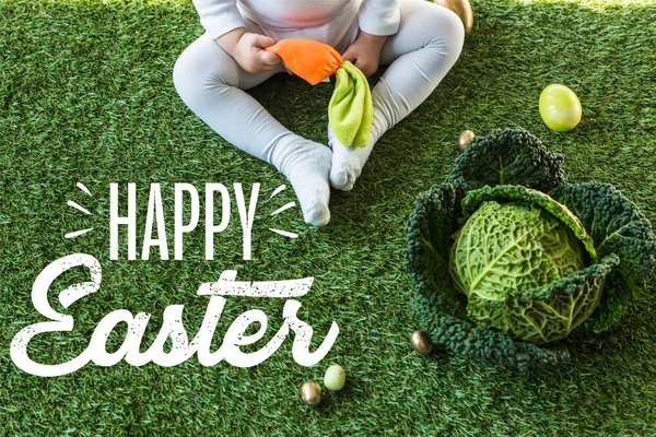 Partial view of child holding toy carrot while sitting near Easter eggs and savoy cabbage on green grass with happy Easter lettering — Stock Photo