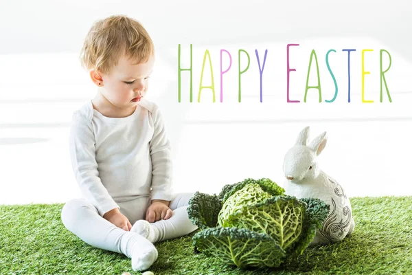 Niño sentado cerca de col de Saboya y conejo decorativo sobre hierba verde con letras de Pascua feliz - foto de stock