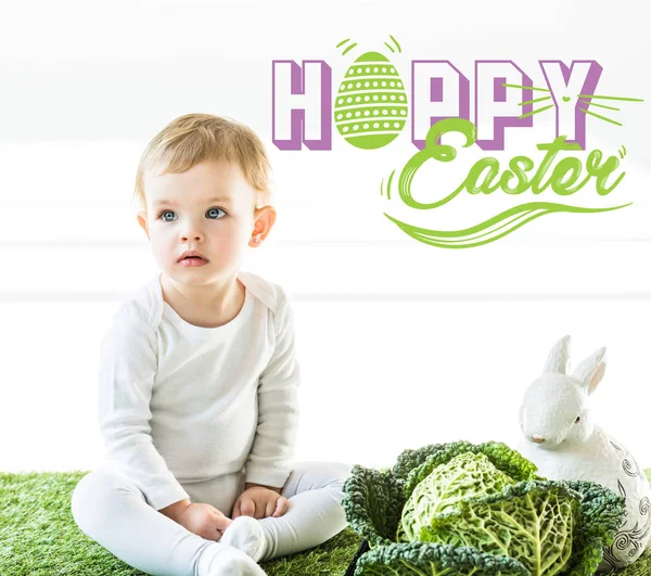 Criança sentada perto de couve lombarda e coelho decorativo na grama verde com letras felizes Páscoa — Fotografia de Stock