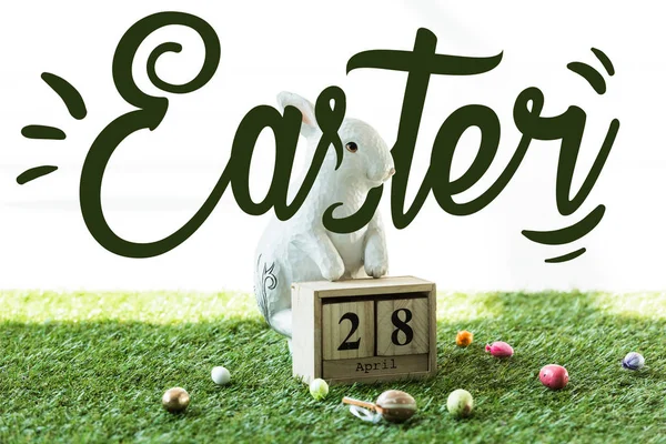 Coelho decorativo, calendário de madeira com data de 28 de abril e ovos de Páscoa coloridos na grama verde com letras de Páscoa — Fotografia de Stock