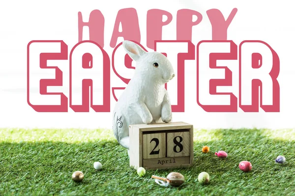 Decorative rabbit, wooden calendar with 28 April date, and colorful Easter eggs on green grass with happy Easter lettering — Stock Photo