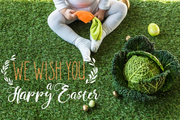 Частичный вид ребенка, держащего игрушечную морковку, сидя рядом с пасхальными яйцами и савойской капусты на зеленой траве с мы желаем вам счастливой Пасхальной надписи — стоковое фото