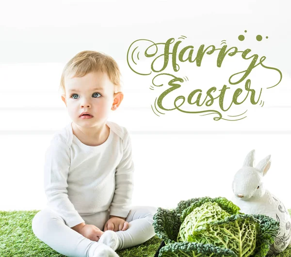 Bambino seduto vicino al verza e coniglietto decorativo su erba verde con scritta verde felice Pasqua — Foto stock
