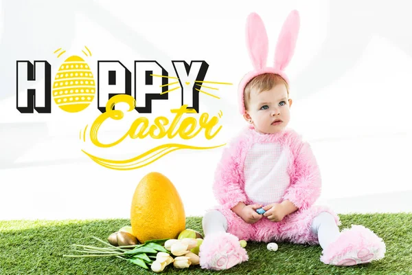 Lindo bebé en traje de conejo sentado cerca de coloridos huevos de pollo, tulipanes y huevo de avestruz amarillo con letras de Pascua feliz - foto de stock