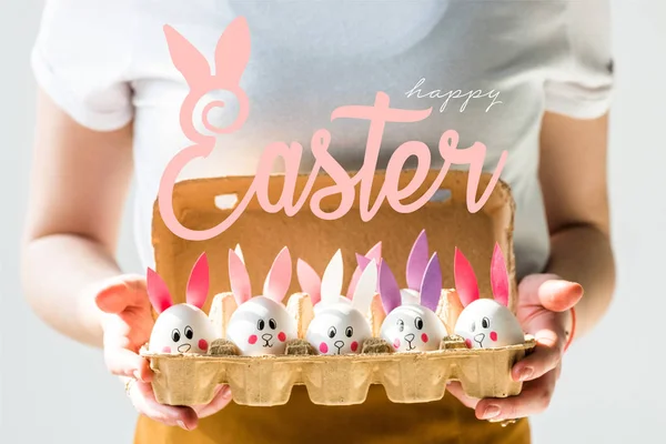 Vista recortada de la mujer sosteniendo bandeja de huevo con huevos de pollo con caras de conejo dibujadas y orejas de papel y letras de Pascua feliz - foto de stock